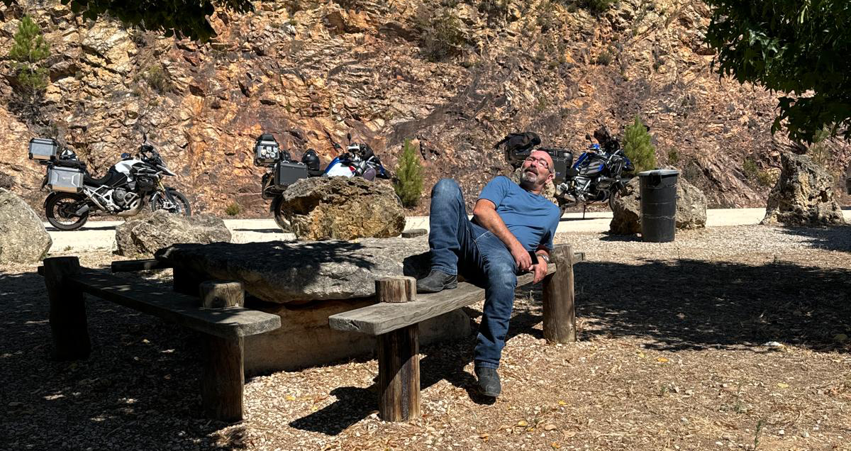 Motorcyclist relaxing on a rustic bench in a scenic rest area with touring bikes parked nearby against a rocky backdrop