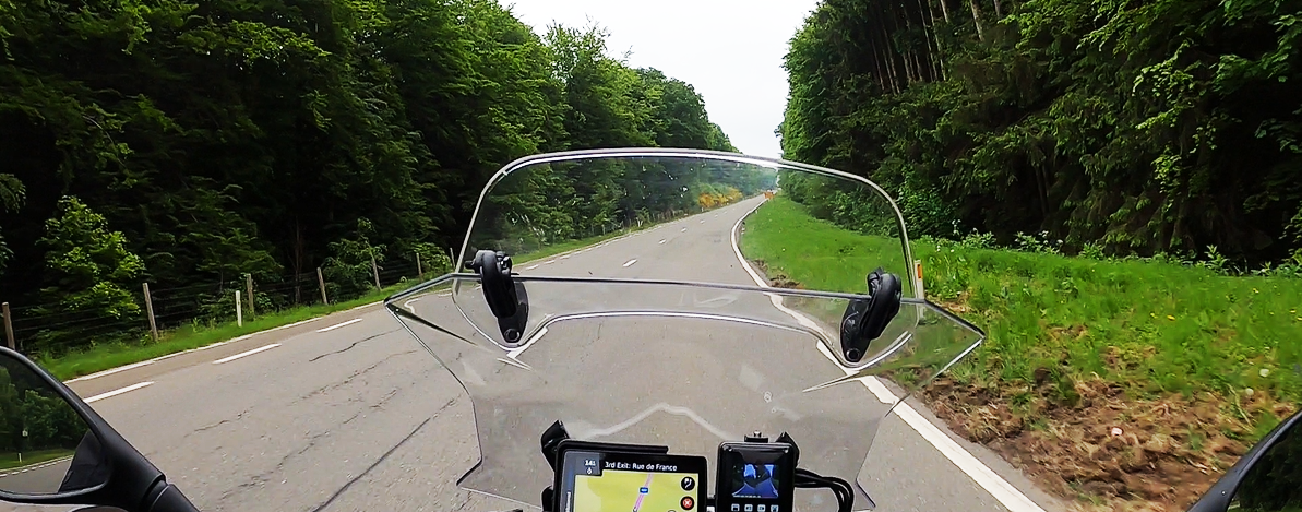 Rider’s view through the windscreen on a forested road in the Black Forest and Vosges region, with GPS navigation visible on the dashboard