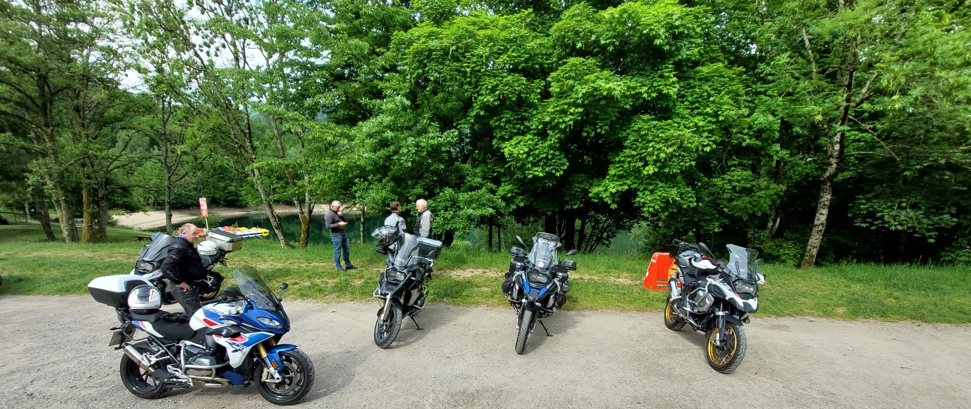 Group of motorcycles parked near a lush green lakeside area in the Vosges Mountains, with riders enjoying a scenic rest stop