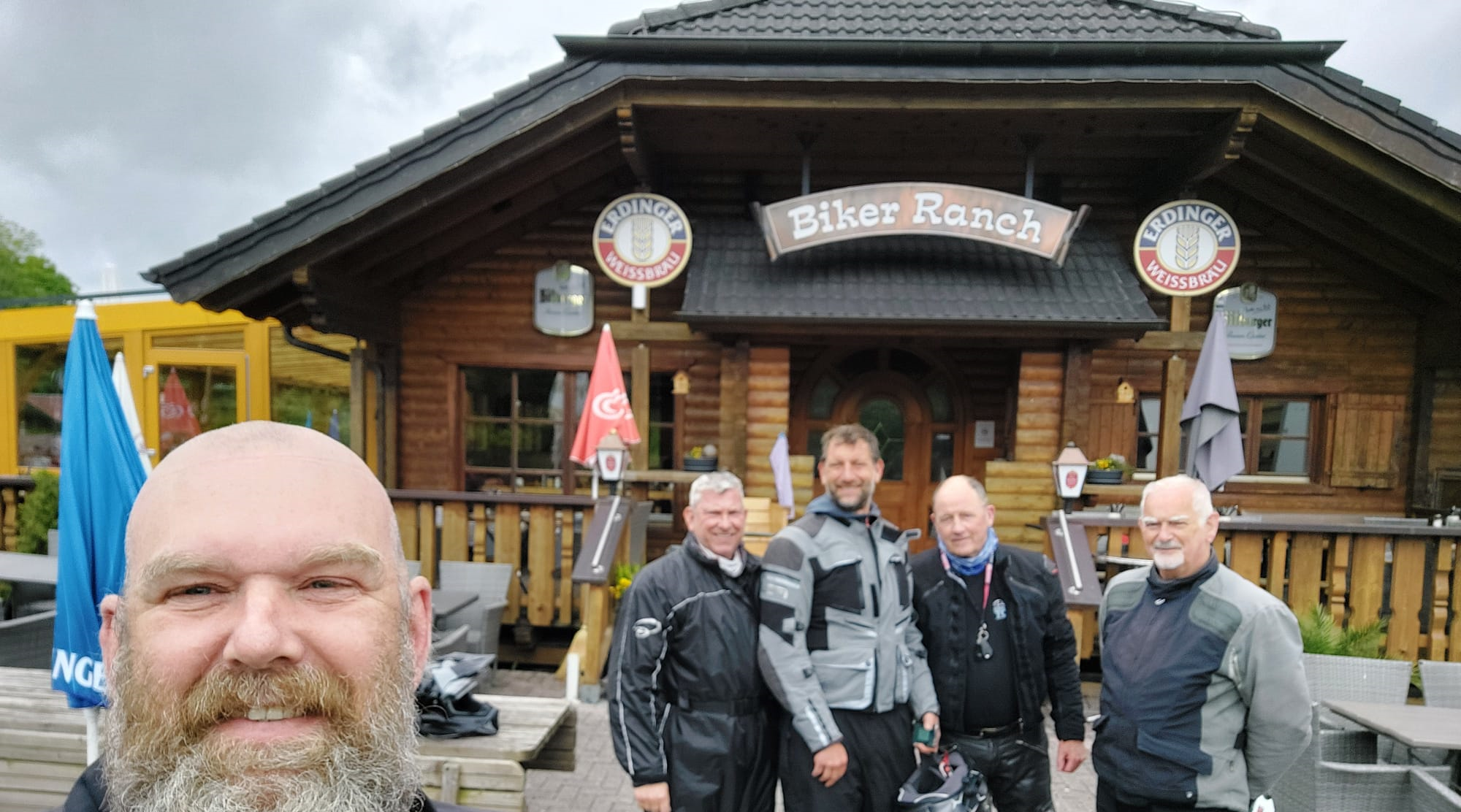 Group of motorcyclists smiling in front of the Biker Ranch, a rustic wooden lodge-style stopover popular with touring riders