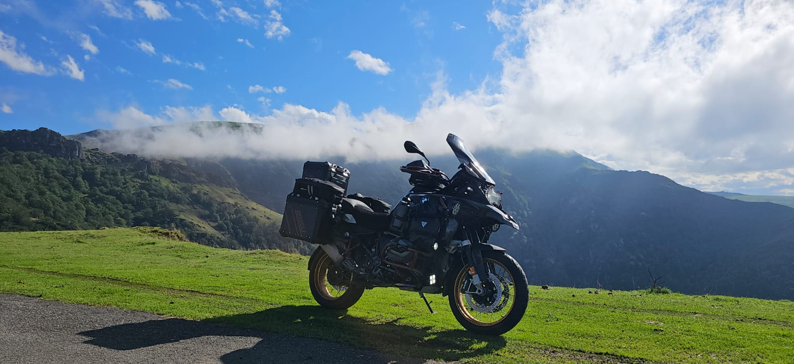 Adventure motorcycle parked on a grassy Pyrenean cliffside with misty mountains and blue skies near Andorra.