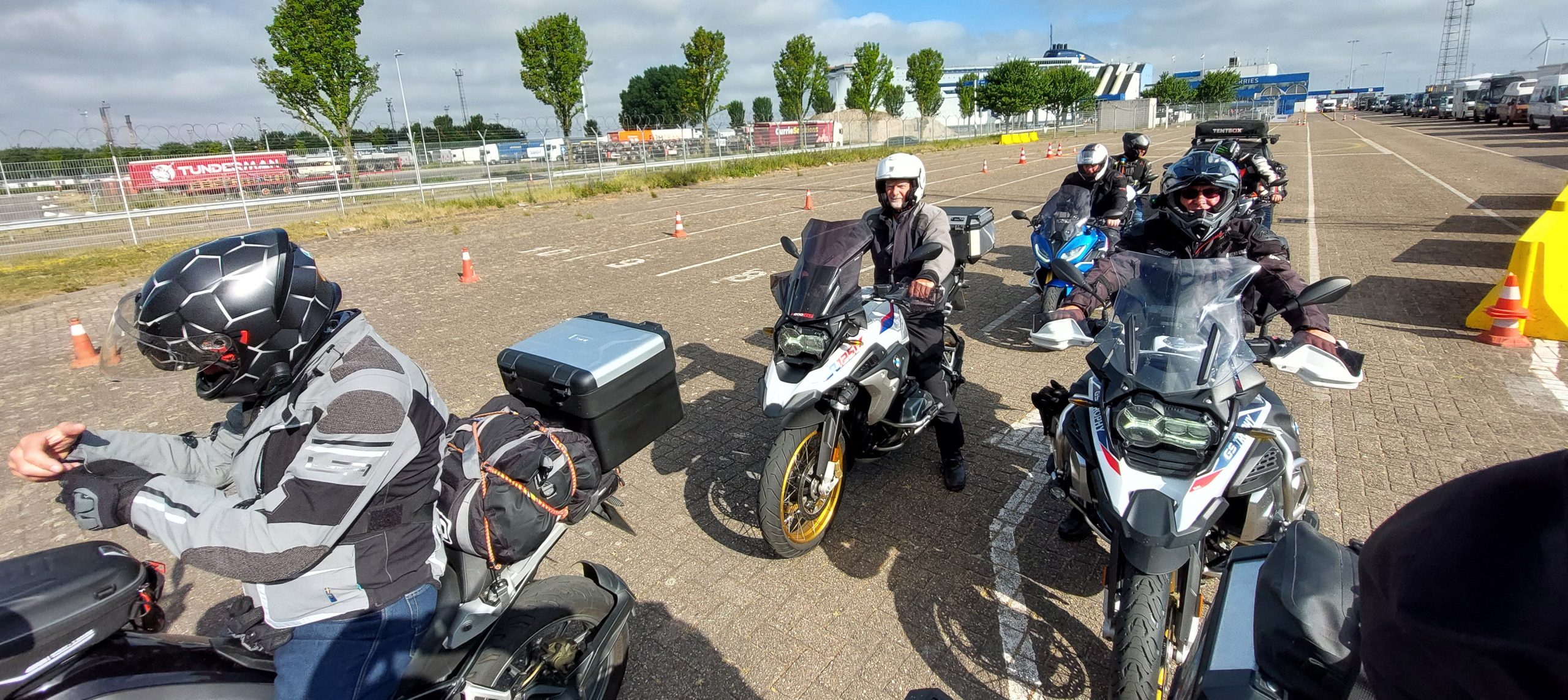 Group of motorcyclists in riding gear lined up on their bikes in a ferry terminal parking area under a bright sky, ready to begin their European motorcycle tour.