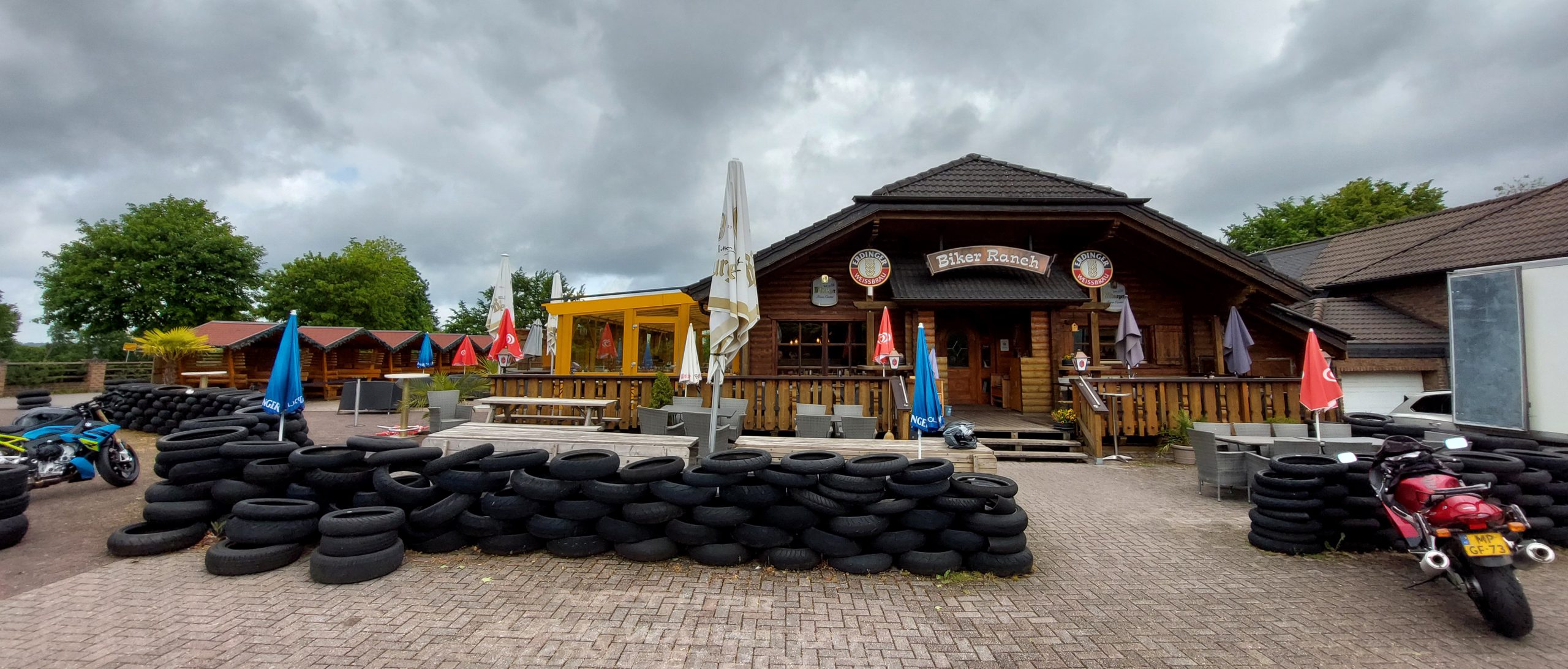 A rustic wooden café called “Biker Ranch” with outdoor seating, surrounded by stacks of tires and motorcycles parked in front, under a cloudy sky.