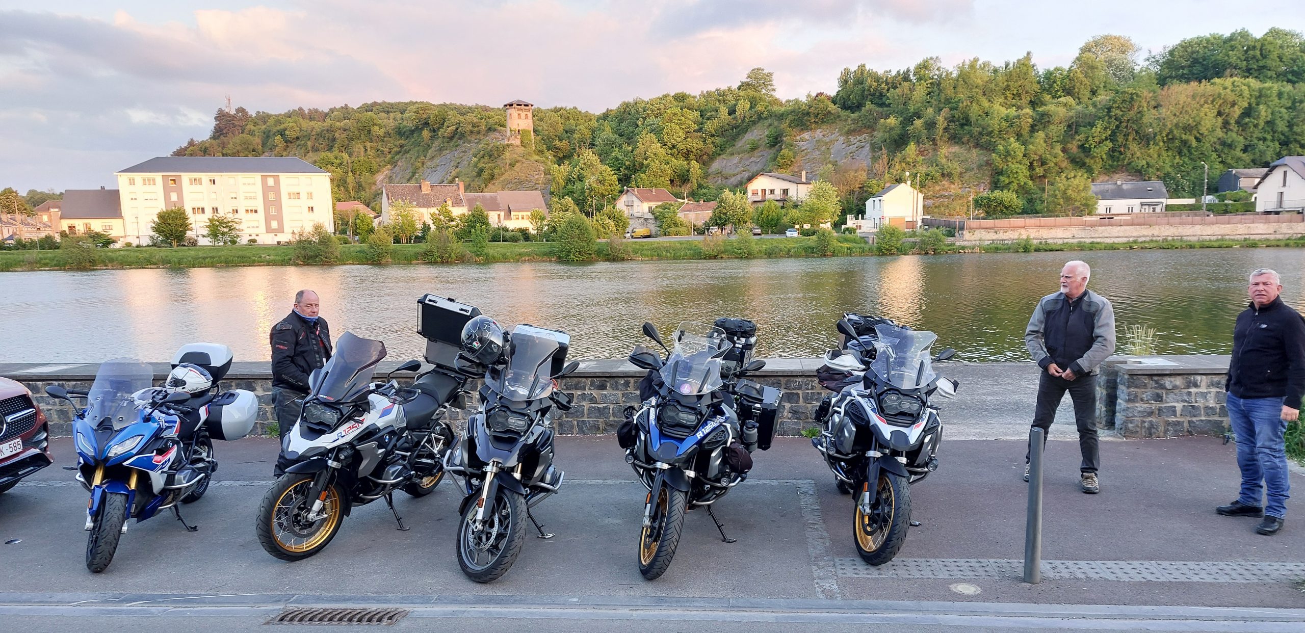 Four motorcyclists standing beside several touring motorcycles parked along the Meuse River in Givet, France, with buildings and green hills in the background.