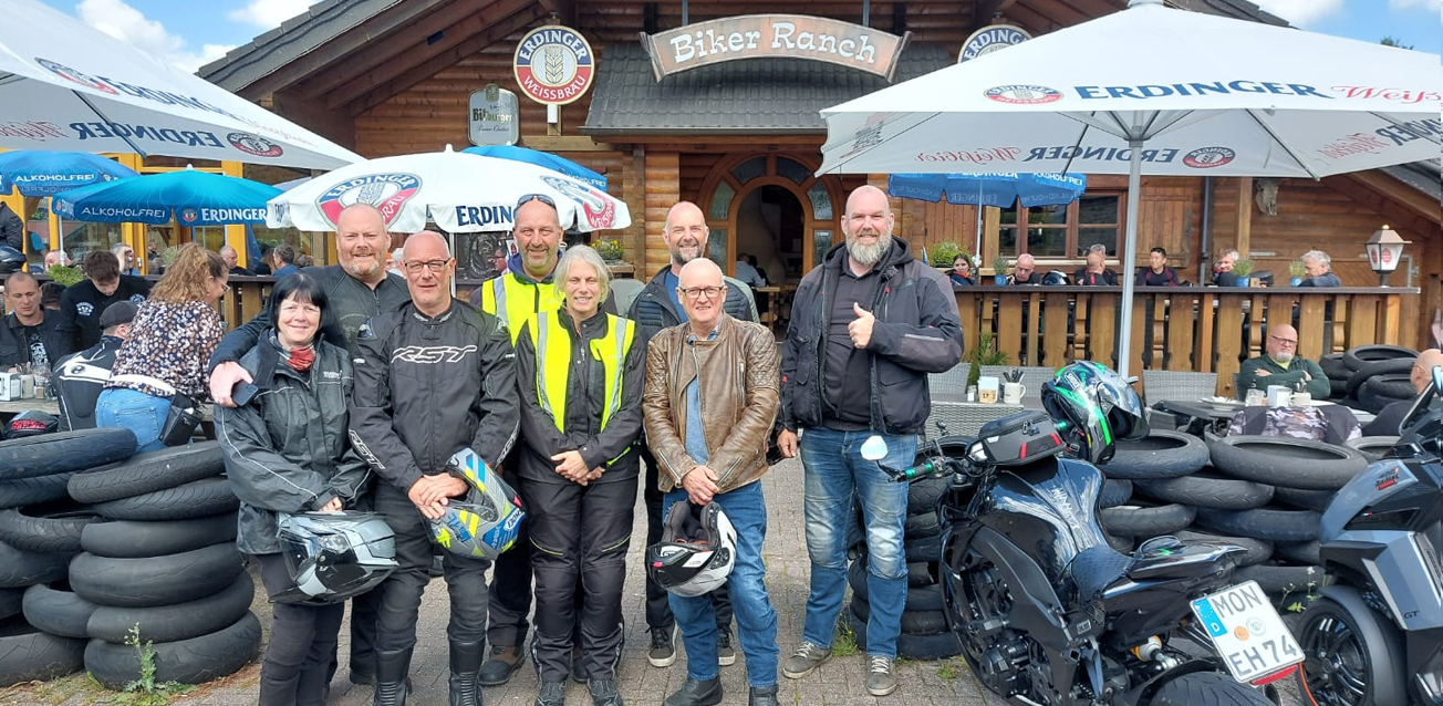 A diverse group of eight motorcycle riders in full riding gear, including leather jackets and high-visibility vests, posing for a photo in front of the "Biker Ranch" cabin. Several riders hold helmets, and a black sport motorcycle is parked in the foreground next to stacks of used tires.