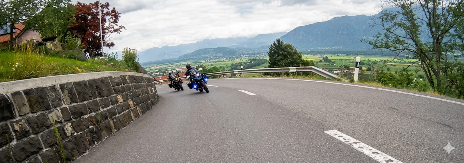 A dynamic, low-angle shot of two motorcyclists leaning into a sharp, sweeping bend on a paved mountain road in the Southern Eifel. To the left is a rustic stone retaining wall, and to the right, a guardrail overlooks a vast green valley and distant mountain ranges under a soft, overcast sky.