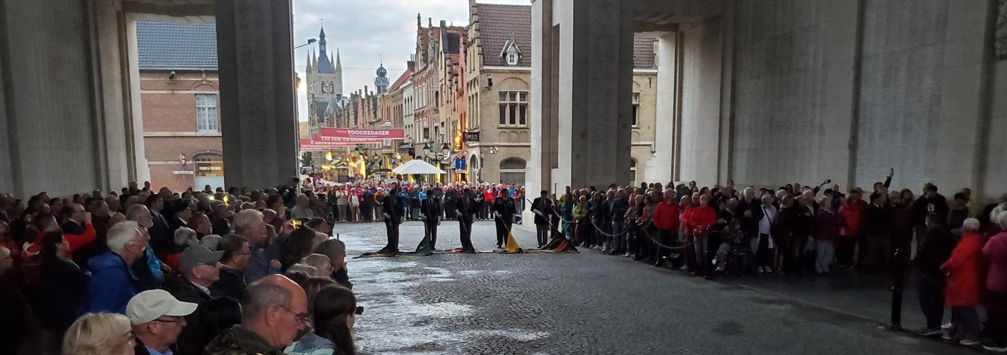 A wide-angle photograph taken from inside the Menin Gate Memorial in Ypres, Belgium, during the Last Post ceremony. A large, respectful crowd of people lines the cobblestone road under the massive stone arches of the gate. In the center, several standard-bearers hold lowered flags. Looking through the archway, the historic buildings and illuminated clock tower of Ypres are visible under a twilight sky.
