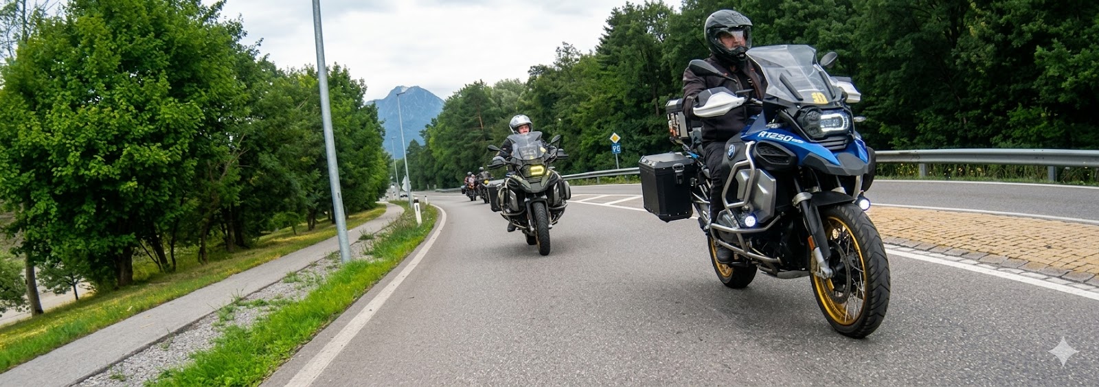 A group of motorcyclists riding in a staggered formation along a scenic, paved road in the Ardennes. The lead rider is on a blue and black BMW R1250 GS adventure bike equipped with panniers. The road is lined with lush green trees under a bright sky, with more riders following behind.