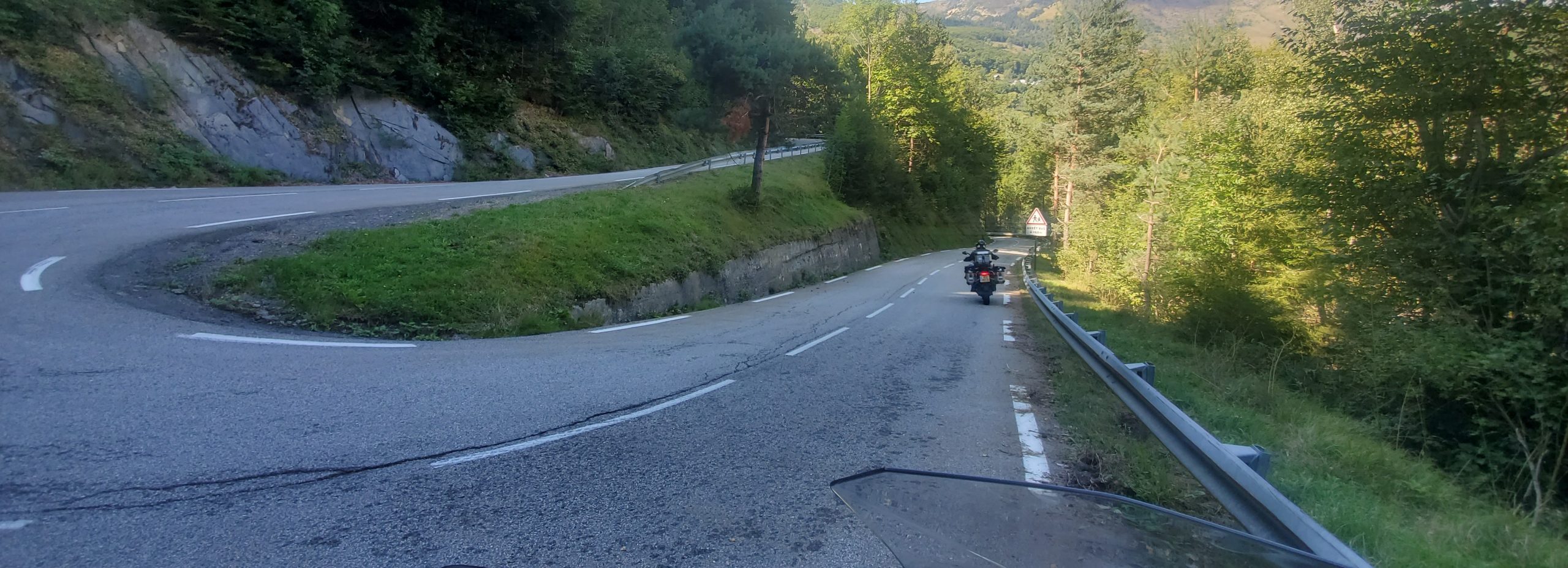 A wide-angle, driver's-perspective photograph from a motorcycle, looking ahead on a winding, paved road (a Col) through a lush, green, mountainous landscape with many trees and a distant valley. The view shows the front of the motorcycle's windshield and the handlebars. A second motorcyclist is visible riding ahead on the road.