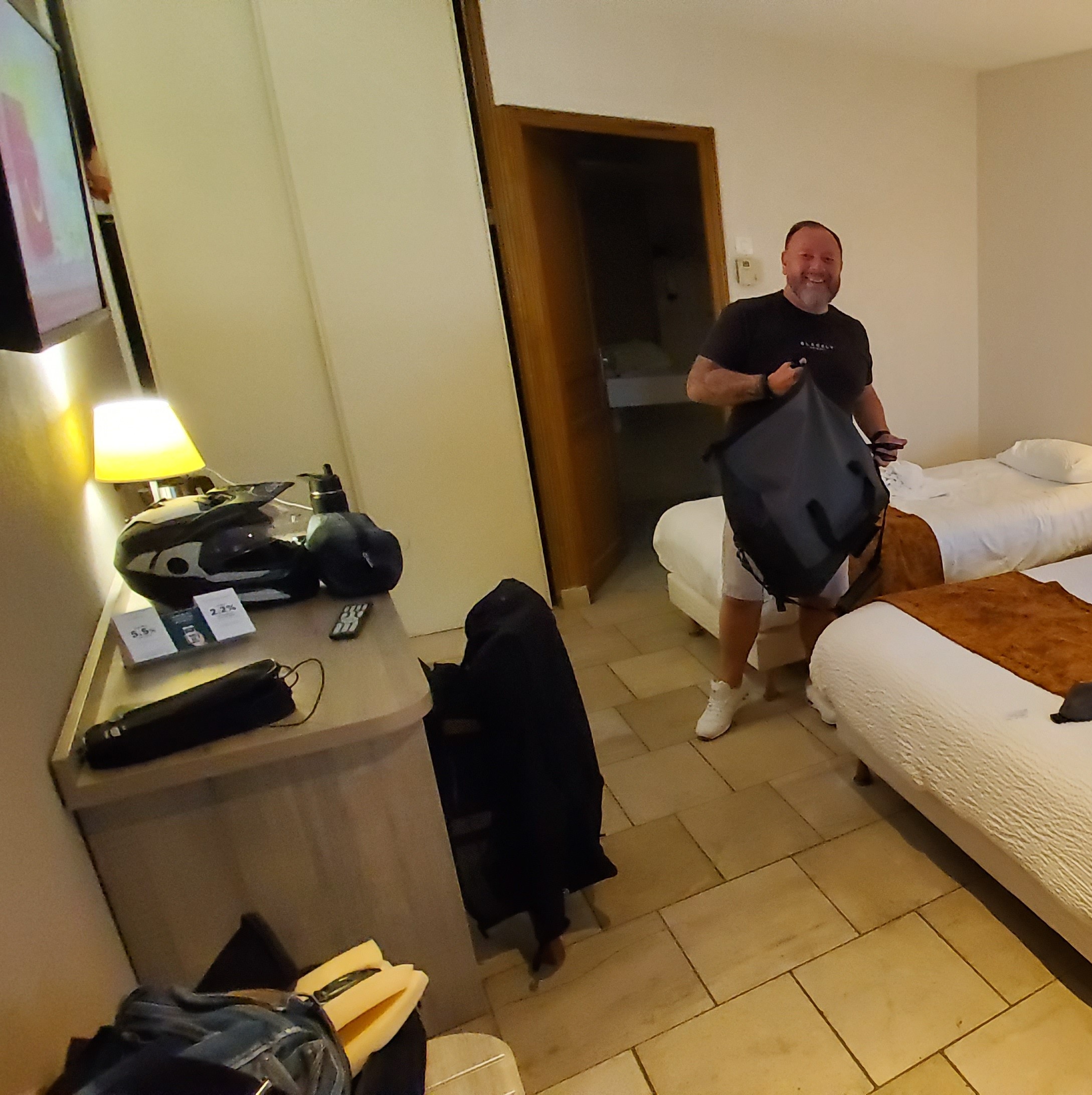 A man with a beard, wearing a dark t-shirt and light shorts, smiles while carrying a large black gear bag in a dimly lit hotel room. A motorcycle helmet and other riding gear are visible on a desk in the foreground.
