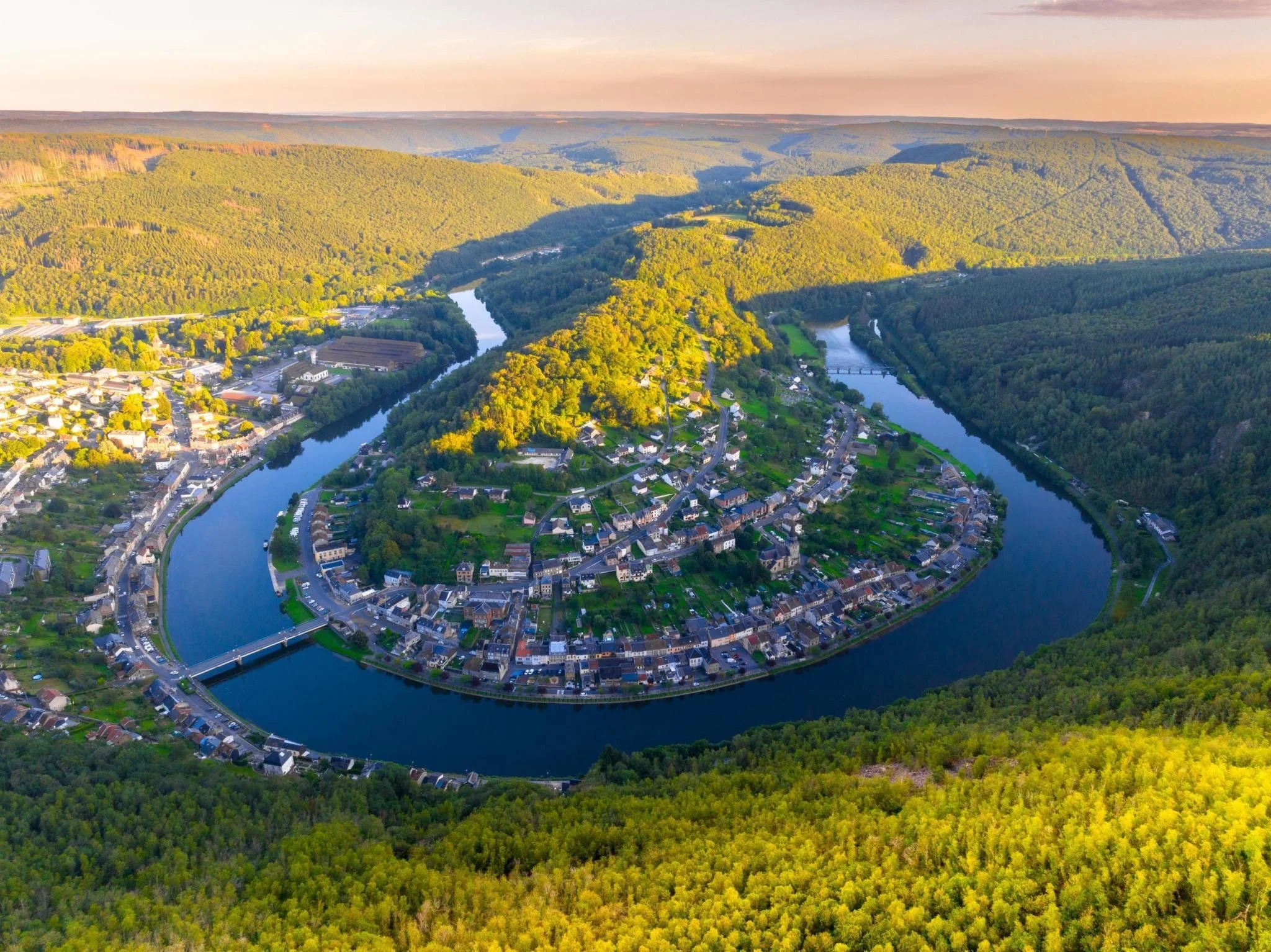 An aerial sunset view of a dramatic U-shaped horseshoe bend in a river, encircling a small town nestled in a deep, forested valley. The lush green hills of the Ardennes region surround the water, with golden light hitting the treetops and a bridge crossing the river in the foreground.