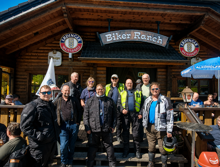 A group of seven motorcyclists in riding gear posing for a photo in front of a rustic wooden building called "Biker Ranch" under a sunny sky.