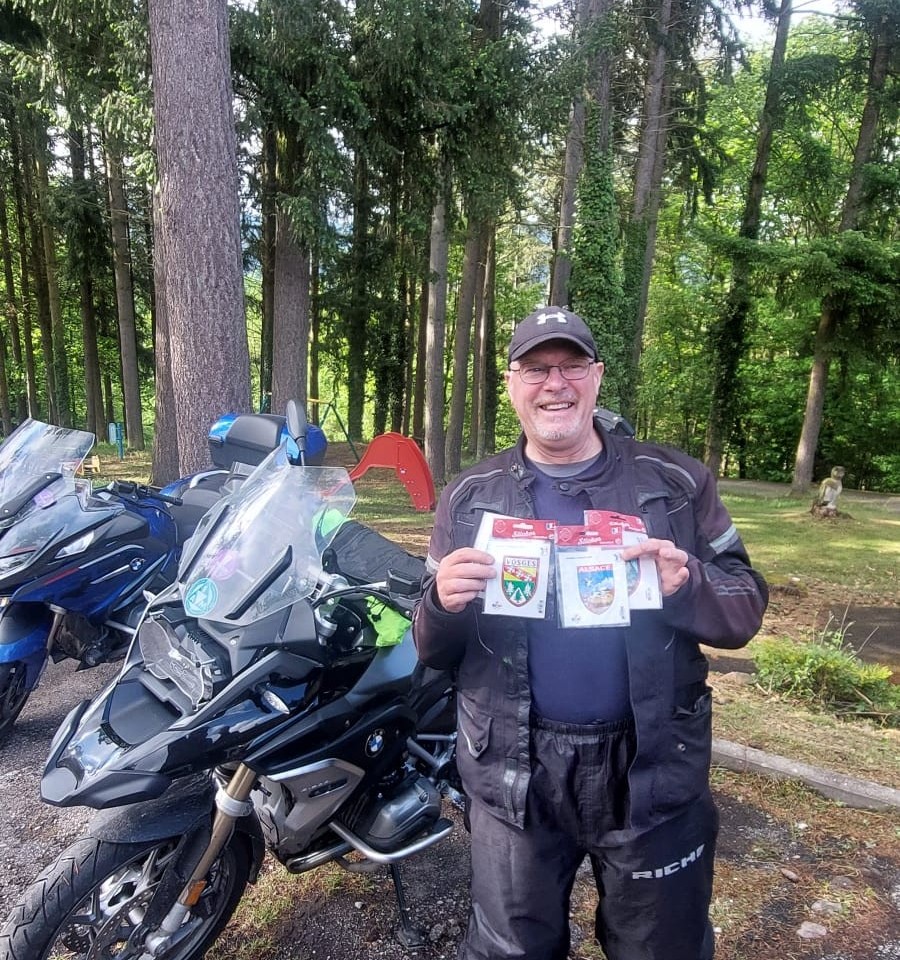A smiling motorcyclist on a Eurobiketours trip stands next to his black BMW adventure motorcycle in a wooded clearing, proudly holding up packaged souvenir stickers from the Vosges and Alsace regions of France.