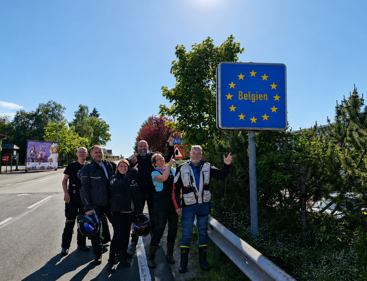 A group of six smiling Eurobiketours motorcycle riders stand on a sunny roadside and point up at a blue European Union border sign reading "Belgien