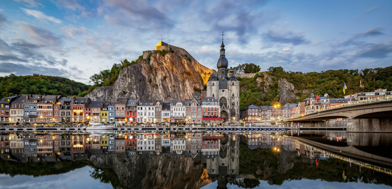A panoramic evening view of Dinant, Belgium, showing the colorful townhouses reflected perfectly in the Meuse River. The Collegiate Church of Our Lady with its iconic pear-shaped bell tower stands in the center, set against a massive limestone cliff topped by the historic Citadelle de Dinant under a dramatic sunset sky.