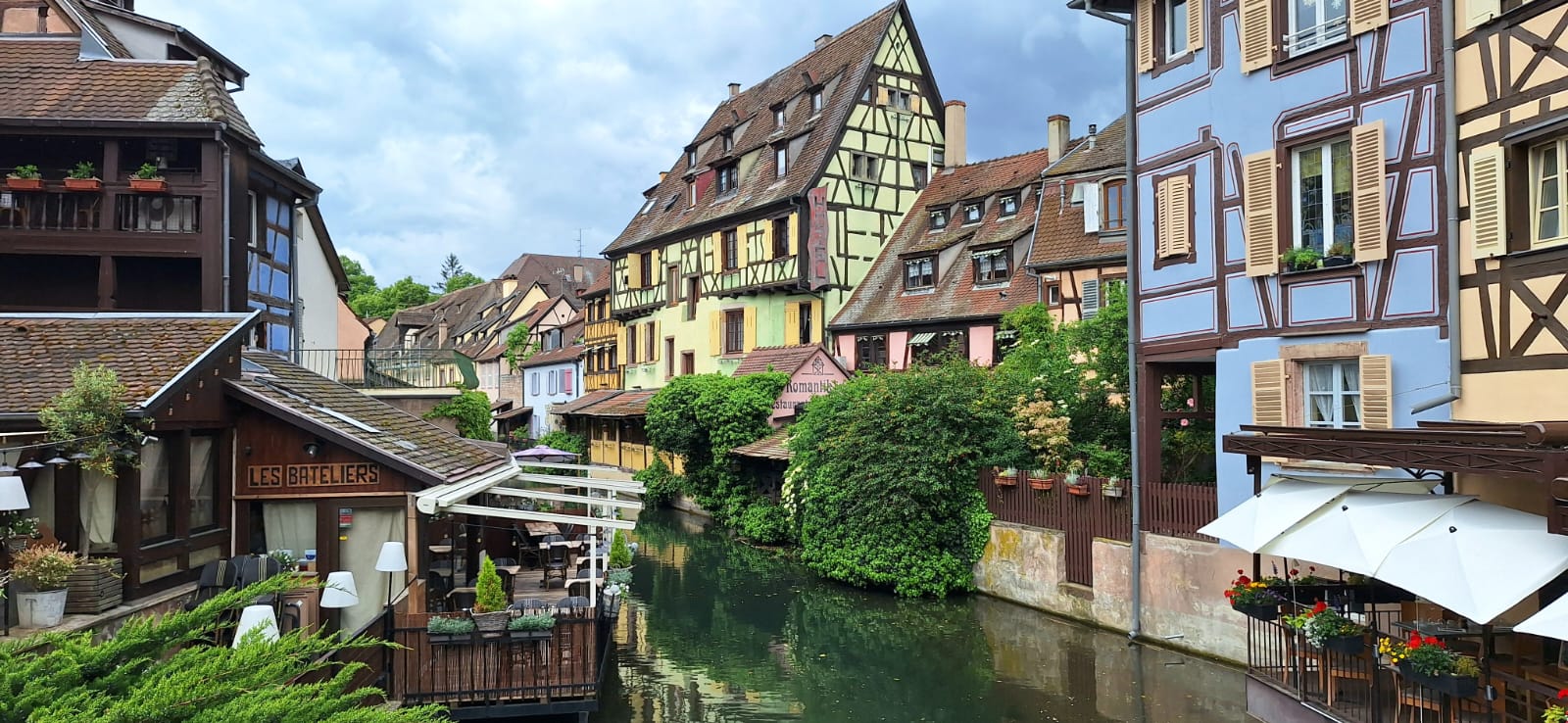 A picturesque view of the canal in the "Little Venice" district of Colmar, lined with historic, colorful half-timbered buildings and outdoor restaurant terraces under a cloudy sky.