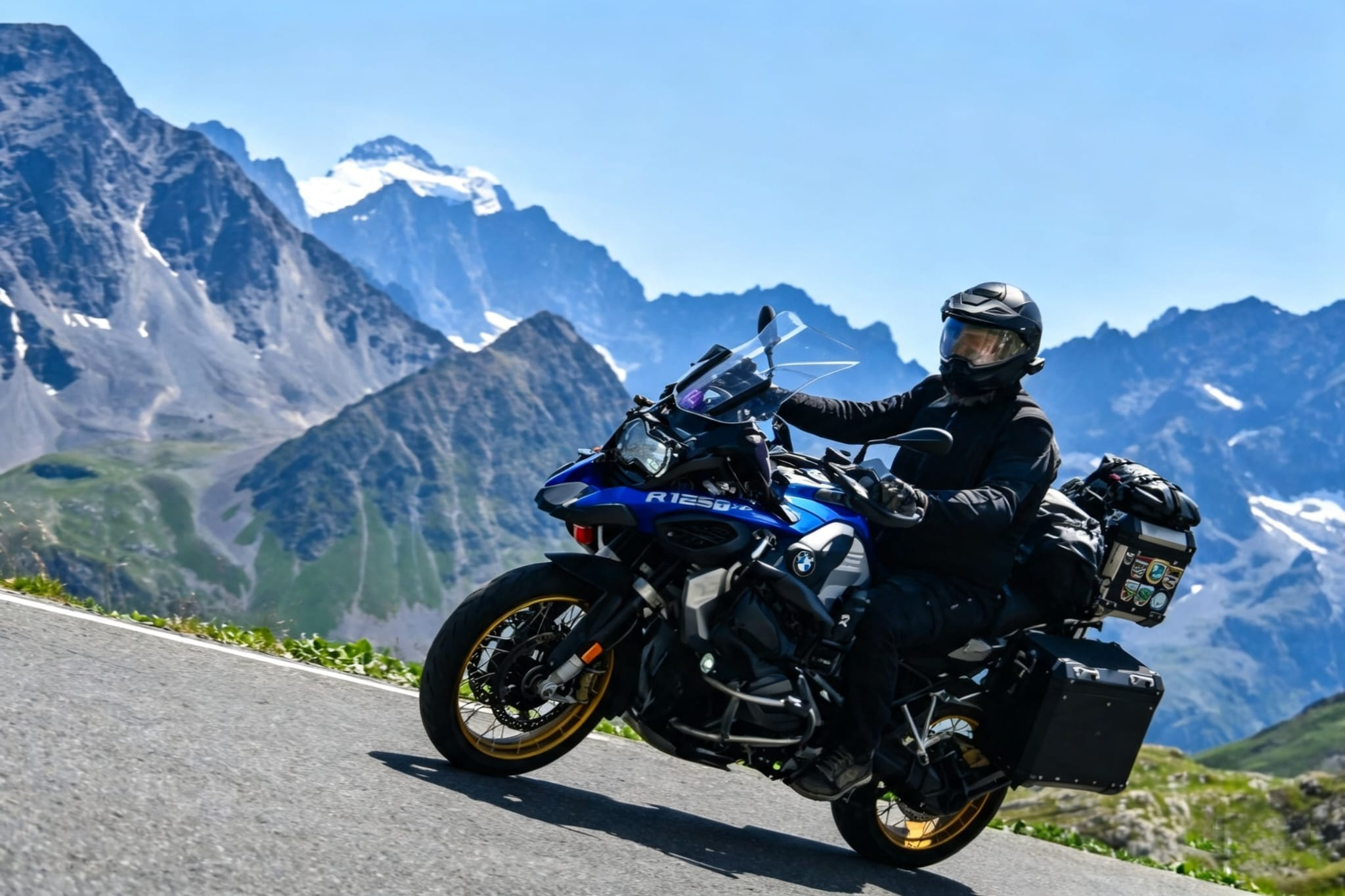 A motorcyclist wearing full black protective gear riding a blue and silver BMW R1250 GS adventure bike loaded with luggage along a mountain road with snow-capped peaks in the background.