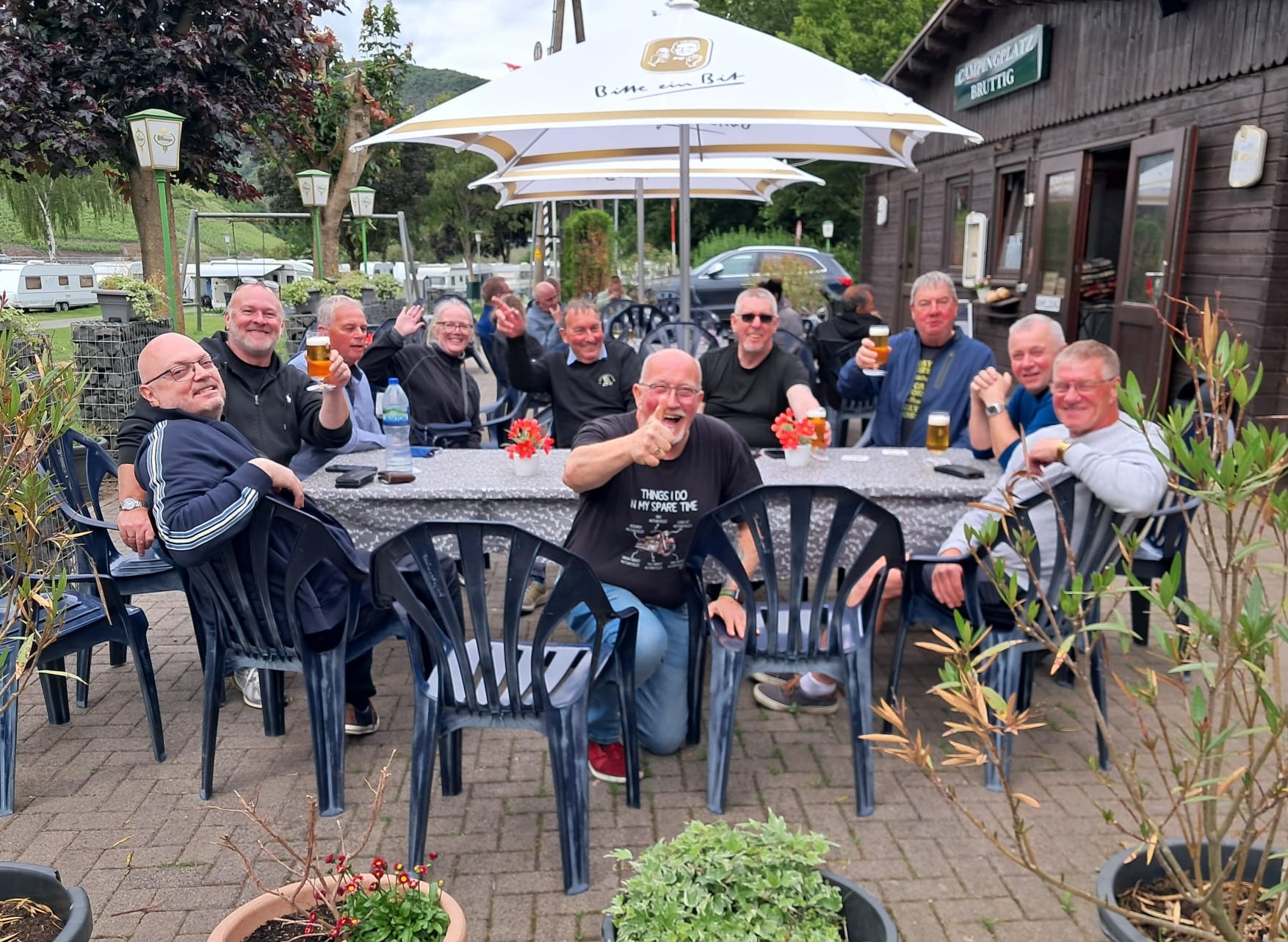 A group of friends sitting together at outdoor patio tables under white umbrellas outside a wooden building marked Campingplatz Bruttig, smiling, waving, and holding up beers during an EU tour.