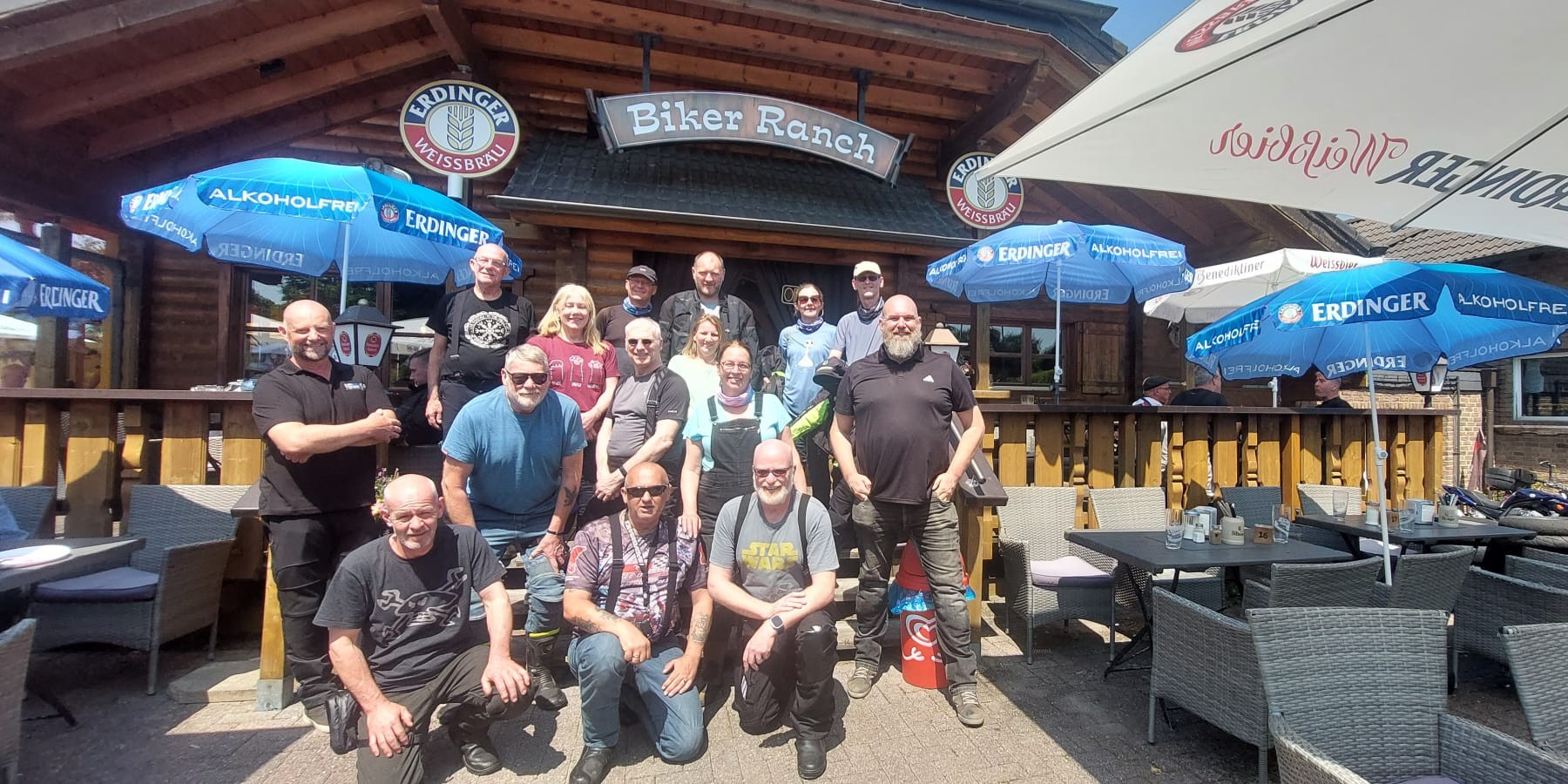 A group of motorcyclists in riding gear posing for a photo in front of a rustic wooden building called "Biker Ranch" under a sunny sky.