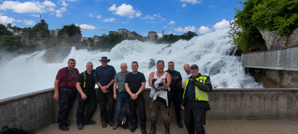 A group of Eurobiketours riders pose together along a concrete viewing platform with a massive, rushing waterfall cascading directly behind them under a blue sky.