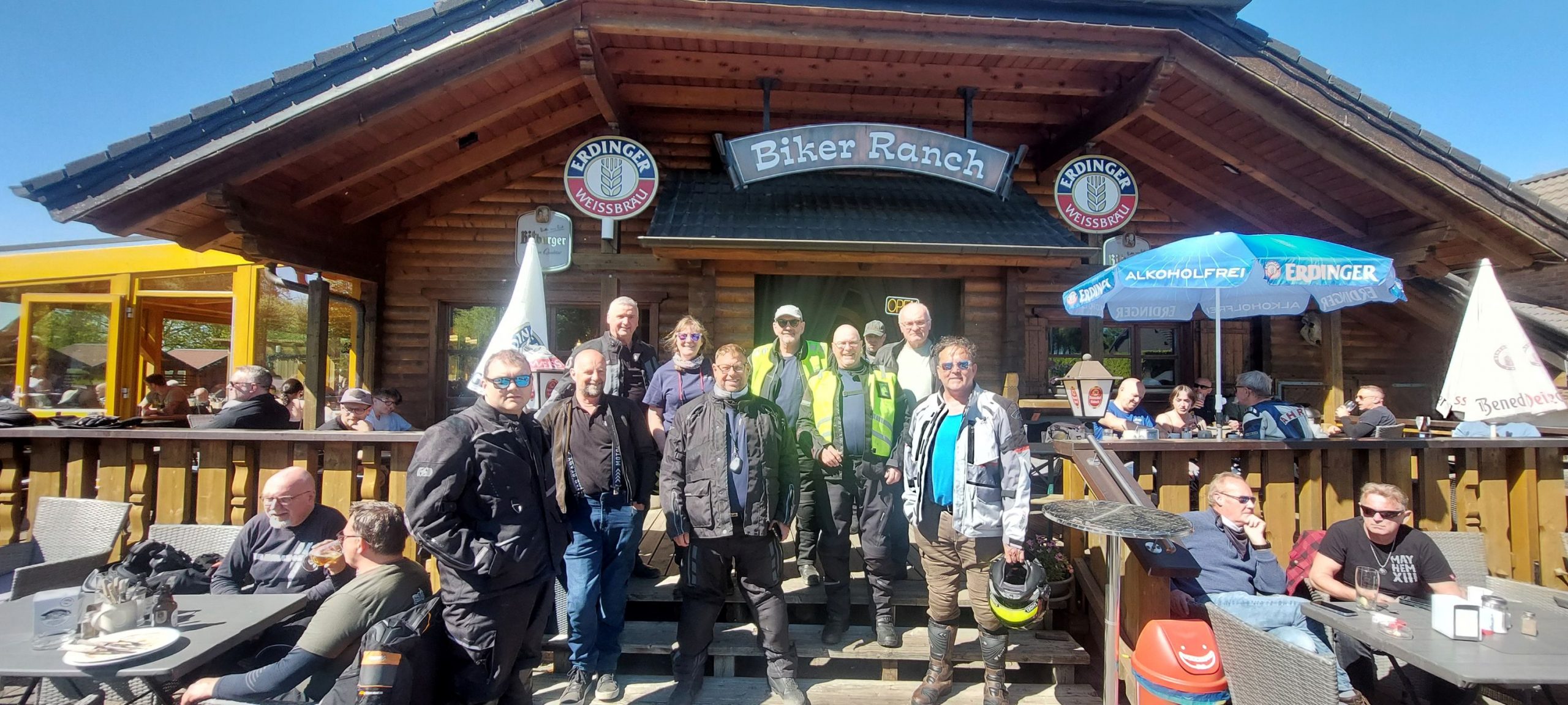 A group of Eurobiketours motorcyclists in riding gear pose for a group photo in front of the "Biker Ranch" log cabin style restaurant, with other patrons sitting at outdoor patio tables on a sunny day.