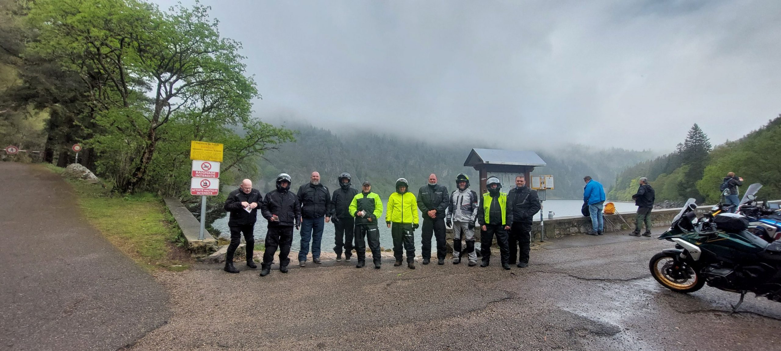 A group of Eurobiketours riders in motorcycle gear stand together on a paved lakeside path during a foggy, overcast day in the Vosges Mountains, with a misty lake and forested hills behind them.