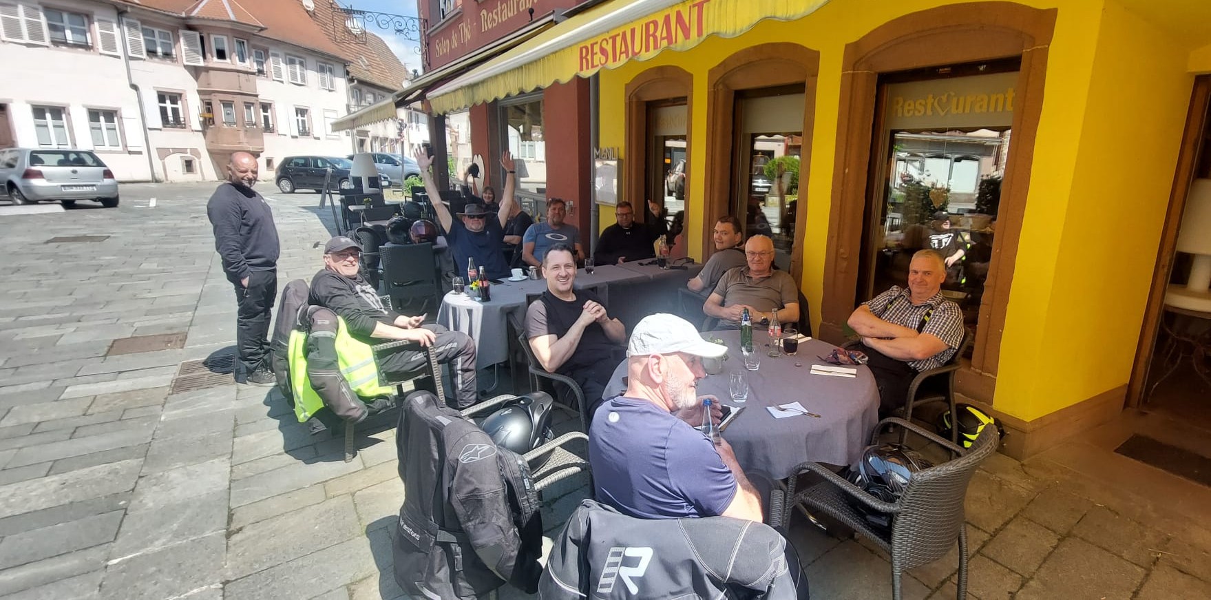 A group of Eurobiketours riders relax and wave cheerfully at outdoor patio tables under the yellow awning of a village restaurant on a sunny day.