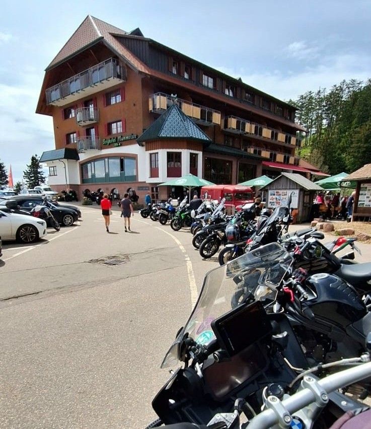 A first-person view from the handlebars of a BMW motorcycle parked in a lot outside the large Berghotel Mummelsee, with numerous other motorcycles parked in a line on a sunny day.